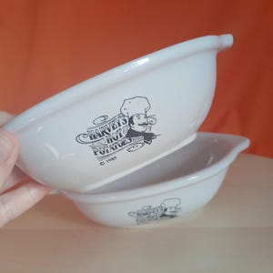 May include: Two white ceramic bowls with a curved design. Each bowl features a black graphic of a chef and the text "Harvey's Hot Potatoes" with the year "1985". The bowls are stacked on top of each other.