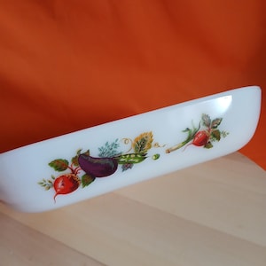 May include: White, oblong serving dish with a colorful vegetable design. The design includes a red radish, a purple eggplant, green peas, and other vegetables. The dish is set against an orange background and a light wood surface.