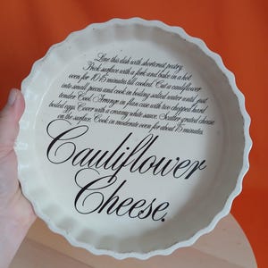 May include: Off-white ceramic pie dish with a scalloped edge. The dish features the words "Cauliflower Cheese" in elegant black script. Recipe instructions are printed around the rim of the dish.