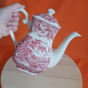 May include: A white and red ceramic teapot with a lid. The teapot has a scenic design with trees and buildings. The handle and spout are white. The lid has a floral pattern. The teapot is held by a hand.