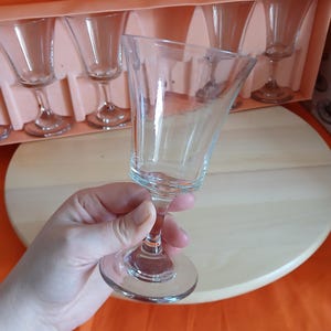 May include: A set of clear glass cordial glasses, with a flared bowl and a slender stem. The glasses are displayed in a peach-coloured box, with one glass held in a hand. The background is a wooden surface and an orange fabric.