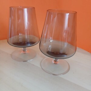 May include: Two clear, stemmed glasses with a brown tint. The glasses have a wide, conical bowl and a circular base. The background is a light wood surface and an orange wall.