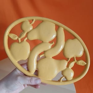 May include: Yellow oval trivet with a fruit design. The trivet features stylized pears, apples, and a banana. The fruit shapes are raised and detailed. The trivet is made of a solid material and has a smooth finish.