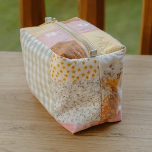 May include: A rectangular quilted pouch featuring a patchwork design of various fabrics, including light green gingham, pink floral prints, yellow checkered patterns, and small orange flowers. The bag has a top zipper closure with a metallic pull.