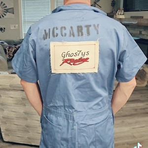 May include: A light blue jumpsuit with the name "MCCARTY" printed across the upper back. Below the name is a rectangular patch with the text "Ghost7ys Racing" in red and white. The jumpsuit has short sleeves and multiple pockets.