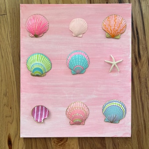 Op de afbeelding: Een roze canvas met een arrangement van kleurrijke schelpenversieringen. De schelpen zijn in verschillende vormen en kleuren, waaronder roze, oranje, blauw en groen. Een witte zeester is ook inbegrepen. De achtergrond is effen roze.