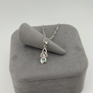 May include: A silver necklace featuring a pendant with a light blue oval gemstone set below a triquetra knot design. The pendant hangs from a twisted silver chain, showcasing a polished metallic finish and a clear, pale blue stone setting.