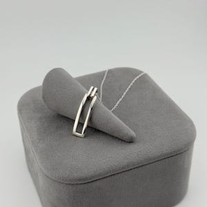 Könnte beinhalten: Eine silberfarbene Anhängerkette mit einem rechteckigen, offenen Rahmendesign. Der Anhänger hängt an einer zarten, dünnen silberfarbenen Kette. Das Metall hat eine glatte, polierte Oberfläche, die das Licht reflektiert und schlicht wirkt.