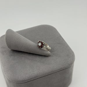 May include: A silver ring featuring a central white opal stone surrounded by a cluster of dark red garnet stones, with a band designed to resemble a leaf with etched vein details, displayed on a gray velvet ring cone stand.