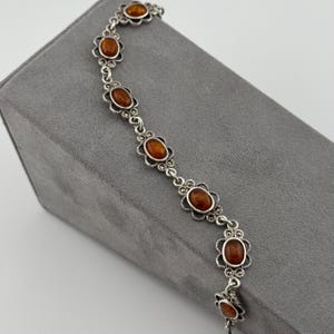 May include: A silver bracelet featuring oval amber-coloured stones set within decorative floral frames. The bracelet is displayed on a grey surface, highlighting its intricate design and warm tones.