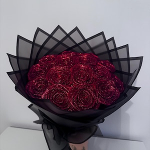 May include: A bouquet of red glitter roses, presented in black paper and sheer black fabric. The roses are densely packed, and the wrapping provides a layered, decorative appearance. The bouquet is held by a person.