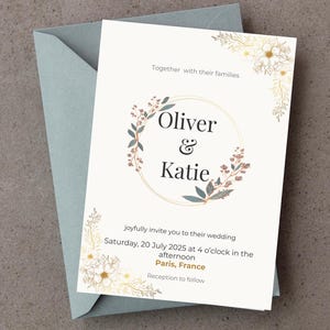 Op de afbeelding: Trouwkaart met de namen "Oliver & Katie" in een bloemenkrans. De uitnodiging staat op een witte kaart met details zoals datum, tijd en locatie in Parijs, Frankrijk, en ligt op een blauwgrijze envelop.