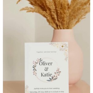 Op de afbeelding: Een trouwkaart met de namen "Oliver & Katie" in een bloemenkransontwerp. De uitnodiging staat op een witte kaart, met details van de trouwdatum, tijd en locatie in Parijs, Frankrijk. Een roze vaas bevat gedroogd siergras.
