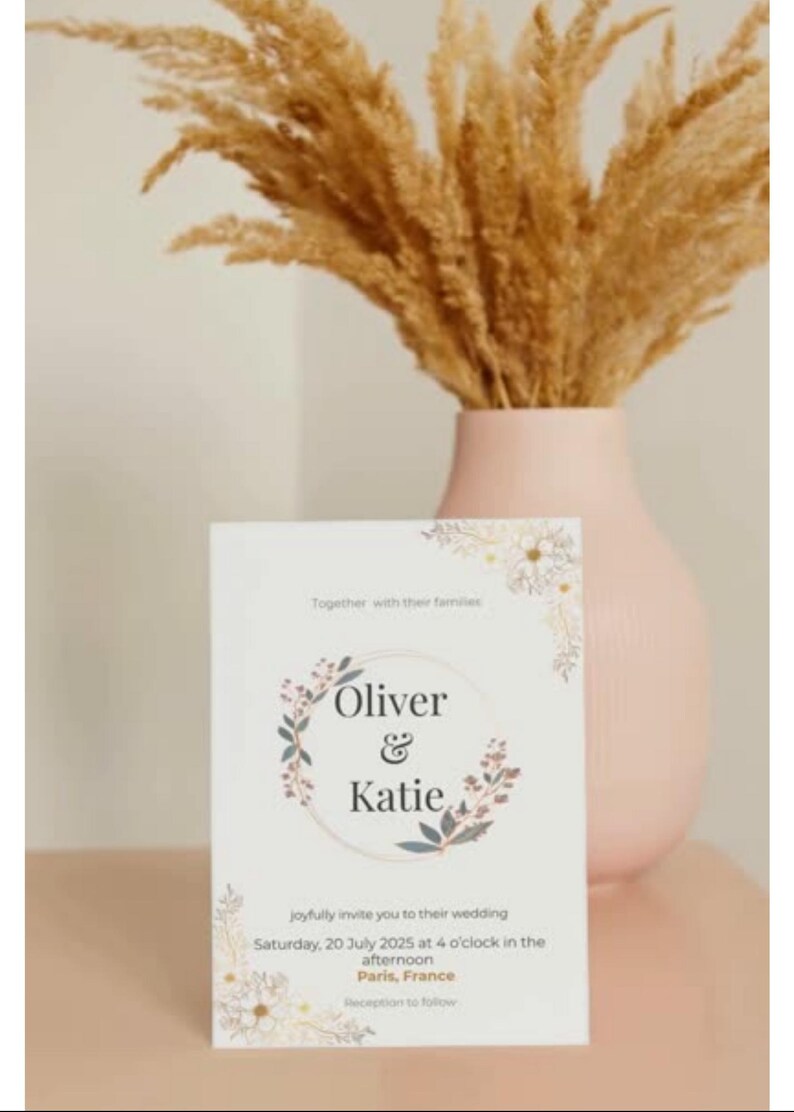 Op de afbeelding: Een trouwkaart met de namen "Oliver & Katie" in een bloemenkransontwerp. De uitnodiging staat op een witte kaart, met details van de trouwdatum, tijd en locatie in Parijs, Frankrijk. Een roze vaas bevat gedroogd siergras.