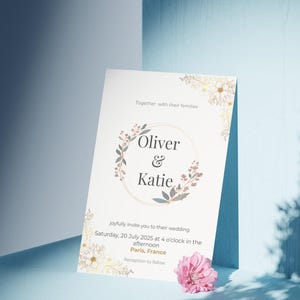 Op de afbeelding: Een huwelijksuitnodiging met de namen "Oliver & Katie" in een bloemenkrans. De uitnodiging bevat de datum, tijd en locatie van de bruiloft in Parijs, Frankrijk. Een roze bloem ligt bij de uitnodiging.