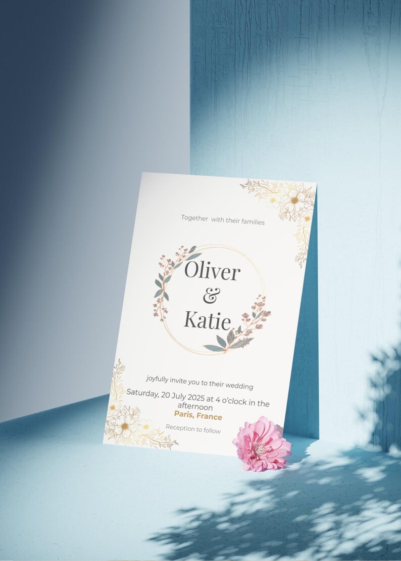 Op de afbeelding: Een huwelijksuitnodiging met de namen "Oliver & Katie" in een bloemenkrans. De uitnodiging bevat de datum, tijd en locatie van de bruiloft in Parijs, Frankrijk. Een roze bloem ligt bij de uitnodiging.