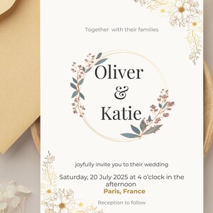 Op de afbeelding: Trouwkaart met de namen "Oliver & Katie" in een bloemenkrans. De uitnodiging vermeldt de datum, tijd en locatie van de bruiloft in Parijs, Frankrijk.