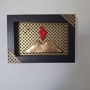 Puede incluir: Obra de arte enmarcada con un marco de madera negro. La obra presenta un fondo de metal dorado con un patrón de cruces. Una forma de montaña de color bronce y un objeto en forma de corazón rojo están en el centro. Acentos dorados en las esquinas.