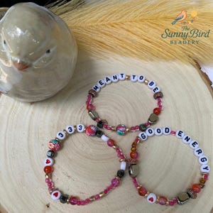May include: Three beaded bracelets with white letter beads spelling "MEANT TO BE", "333", and "GOOD ENERGY". The bracelets feature pink, red, and gold beads, with heart and geometric accents. The Sunny Bird Beadery logo is visible.