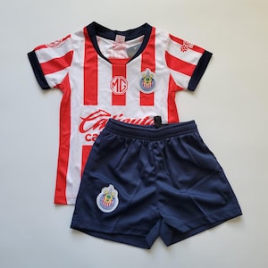 May include: A red and white striped soccer jersey and navy blue shorts set. The jersey has a round neck, short sleeves, and a team logo on the chest. The shorts have an elastic waistband and a matching team logo.