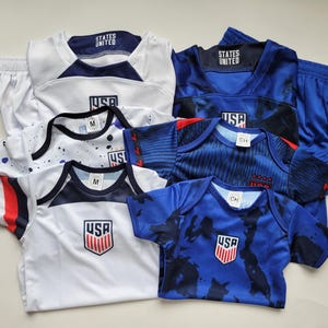 May include: A collection of athletic apparel including white and blue jerseys and onesies featuring the text STATES UNITED and USA logos. The garments include short-sleeved shirts, a white pair of shorts, and a blue pair of shorts with a drawstring.