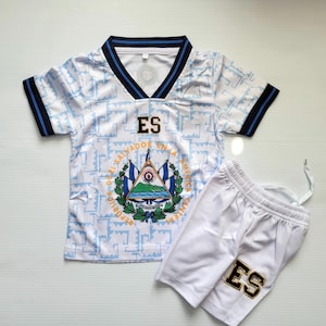 May include: A white and light blue soccer jersey and shorts set. The jersey has a V-neck with navy blue trim and the letters "ES" on the chest. The shorts also have "ES" on the leg. The set features the El Salvador coat of arms.