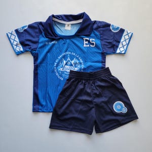 May include: A blue and navy soccer uniform set. The shirt has a collar and the letters "ES" on the front. The shorts have an emblem on the leg. The shirt has the text "EL SALVADOR" and "REPUBLICA DE EL SALVADOR EN LA AMERICA CENTRAL".