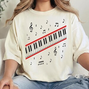 May include: Cream-coloured t-shirt with a piano keyboard design, red border, and musical notes. The keyboard is black and white, with treble clef and other musical symbols scattered around. A casual, music-themed apparel item.