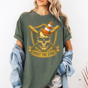 May include: Olive green t-shirt featuring a skull wearing a traffic cone, crossed baseball bats, and the text "HOIST THE CONE" in a gold color. The design is on a shield shape.