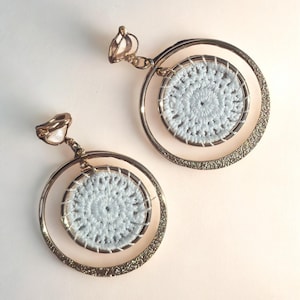 May include: A pair of gold-toned earrings with a circular design. Each earring features a light blue crocheted circle within a larger gold ring. The outer ring has a textured, glittery appearance, and the earrings have a clip-on style.