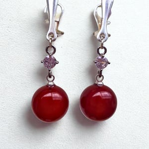 May include: A pair of clip-on earrings featuring a silver-tone clip, a small light purple gemstone, and a large, round, glossy red bead. The earrings are displayed against a white background.