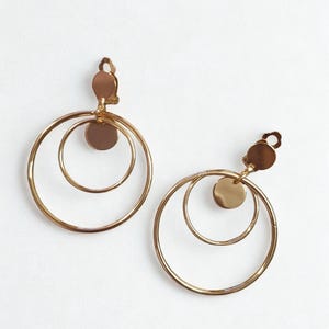 May include: A pair of gold-colored clip-on earrings. Each earring features three concentric hoops and a small, circular accent. The earrings have a shiny finish and are set against a white background.