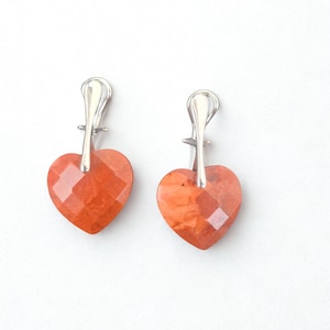 May include: A pair of heart-shaped earrings. Each earring features a faceted, reddish-orange gemstone heart suspended from a silver-colored metal hook. The earrings are displayed against a white background.