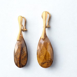 May include: A pair of gold-colored earrings with teardrop-shaped tiger's eye gemstones. The gemstones are faceted, displaying a mix of brown and gold hues. The earrings have a simple, elegant design.