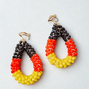 May include: A pair of teardrop-shaped earrings with a gold-colored clip-on closure. The earrings are made of small beads in black, red, and yellow, resembling a flag. The earrings are on a white background.
