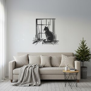 May include: A black metal cat silhouette wall art piece. The cat is sitting in front of a window with a minimalist design. The artwork is mounted on a white wall above a beige sofa with pillows and a blanket. A small side table and a Christmas tree are also in the room.