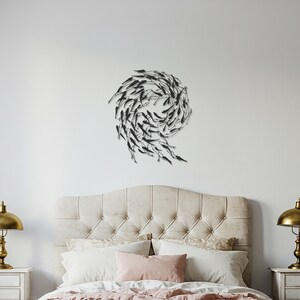 May include: Black metal wall art depicting a school of fish in a swirling pattern. The artwork is mounted on a white wall above a bed with a tufted headboard, pillows, and a pink comforter. Two gold lamps flank the bed.
