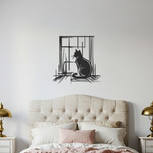 May include: A black metal wall art piece depicting a cat silhouette seated in front of a window. The cat is facing to the left, with a detailed outline. The window frame and surrounding lines add a sketched effect. The artwork is mounted on a white wall above a bed.