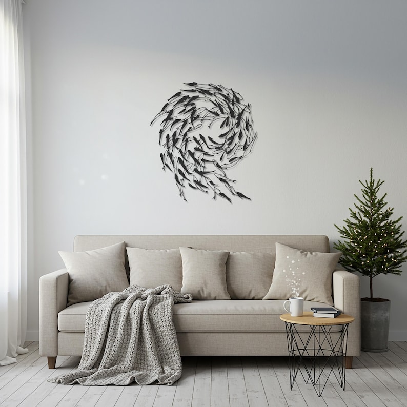 May include: A living room scene featuring a beige sofa with throw pillows and a gray blanket. A black metal sculpture of fish in a spiral formation hangs on the white wall. A small side table and a potted Christmas tree complete the decor.