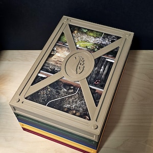 Op de afbeelding: Een rechthoekige bordspeldoos met een beige kunststof frame en een centraal rond embleem. Het deksel toont een bedrukte scène van een stenen pad, houten planken en kasseien. De doos is gestapeld op groene, blauwe, gele en rode lagen.