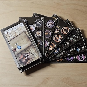 Peut inclure: Un jeu de société avec une boîte de cartes et plusieurs plateaux noirs remplis de jetons circulaires. Les cartes ont un design vintage avec du texte et des illustrations. Les jetons présentent des portraits de personnages de différentes couleurs.