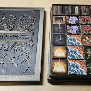 以下が含まれることがあります： 「ELEMENTS」の文字が銀色のフレームに入った灰色のボードゲームボックス。ボックスは開いており、ゲームの駒、カード、トークンが表示されています。カードには、火、水、その他の要素をテーマにした画像が描かれています。ゲームの駒は黒と紫です。