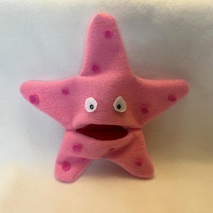 May include: A pink starfish puppet with a wide open mouth and white eyes. The puppet has dark pink spots and is made of a soft material. The starfish is a fun toy for kids.
