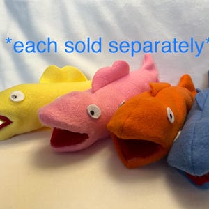 May include: Four colorful felt fish puppets in yellow, pink, orange, and blue. Each puppet has a wide open mouth with a red interior and white eyes. The text "each sold separately" is above the puppets.