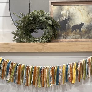 May include: A decorative garland with colorful fabric strips in shades of yellow, blue, orange, and green hangs on a wooden mantel. The mantel also features a framed picture of bears, candles, and a wreath.