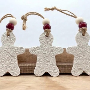 May include: Three white ceramic gingerbread man ornaments with embossed floral patterns. Each ornament has a jute string, a white bead, and a red bead for hanging. The ornaments are displayed on a wooden surface.