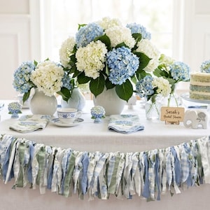 May include: A beautifully decorated table setting with white and blue hydrangeas in vases, teacups, napkins, and a cake. A garland of fabric strips in blue and green adorns the table's edge. A wooden sign reads "Sarah's Bridal Shower."