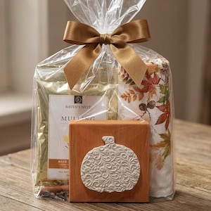 May include: A gift set in a clear plastic bag tied with a brown satin ribbon containing a gold-toned package labeled Raven's Nest Mulling, a white towel with an autumn leaf pattern, and a square wooden block featuring a white embossed pumpkin.