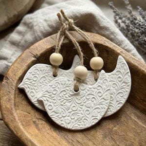 May include: Three white bird-shaped ornaments featuring an intricate embossed floral pattern. Each ornament is suspended by a tan jute twine loop accented with a single smooth, round, light-colored wooden bead at the top of the bird figure.
