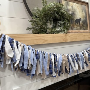 May include: A decorative garland made of fabric strips in shades of blue, white, and tan. The garland is draped across a wooden mantel, with a painting of a bear and a green wreath in the background. The garland adds a rustic touch to the decor.
